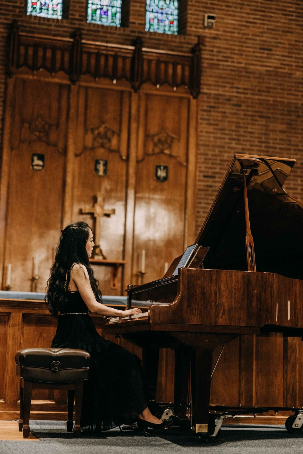Woman in black dress playing grand piano in elegant wood-paneled church with stained glass windows.