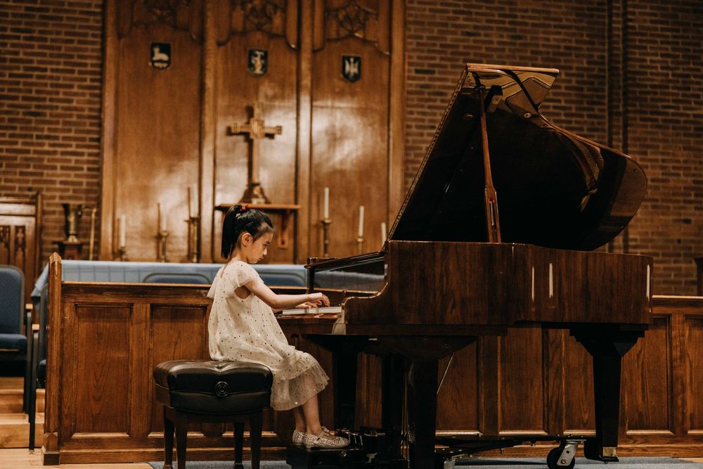 Young girl in white dress playing grand piano in elegant wood-paneled church with cross backdrop.