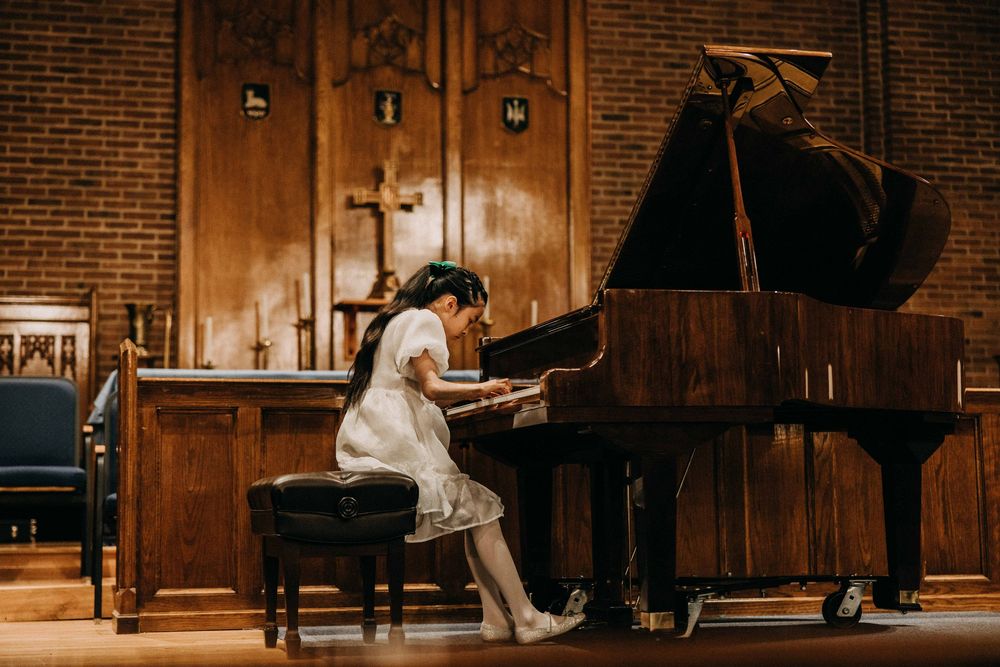 Young girl in white dress playing grand piano on stage in elegant wood-paneled church hall during recital.