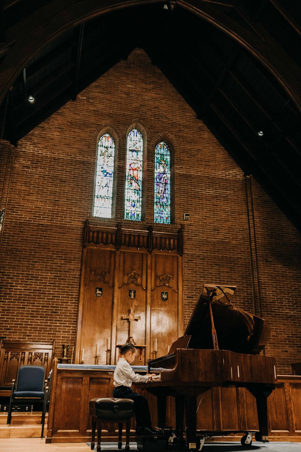 Young pianist performs on grand piano in elegant brick church with stunning stained glass windows and wooden altar backdrop.