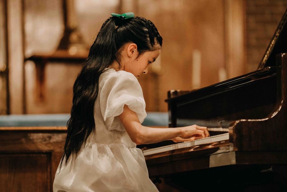 Young girl in white dress playing grand piano, focused on keys, long dark hair with green bow.