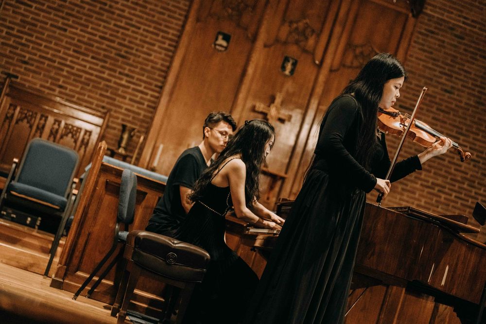 Three musicians perform chamber music, with a violinist and pianist in elegant black attire inside a wood-paneled concert hall.