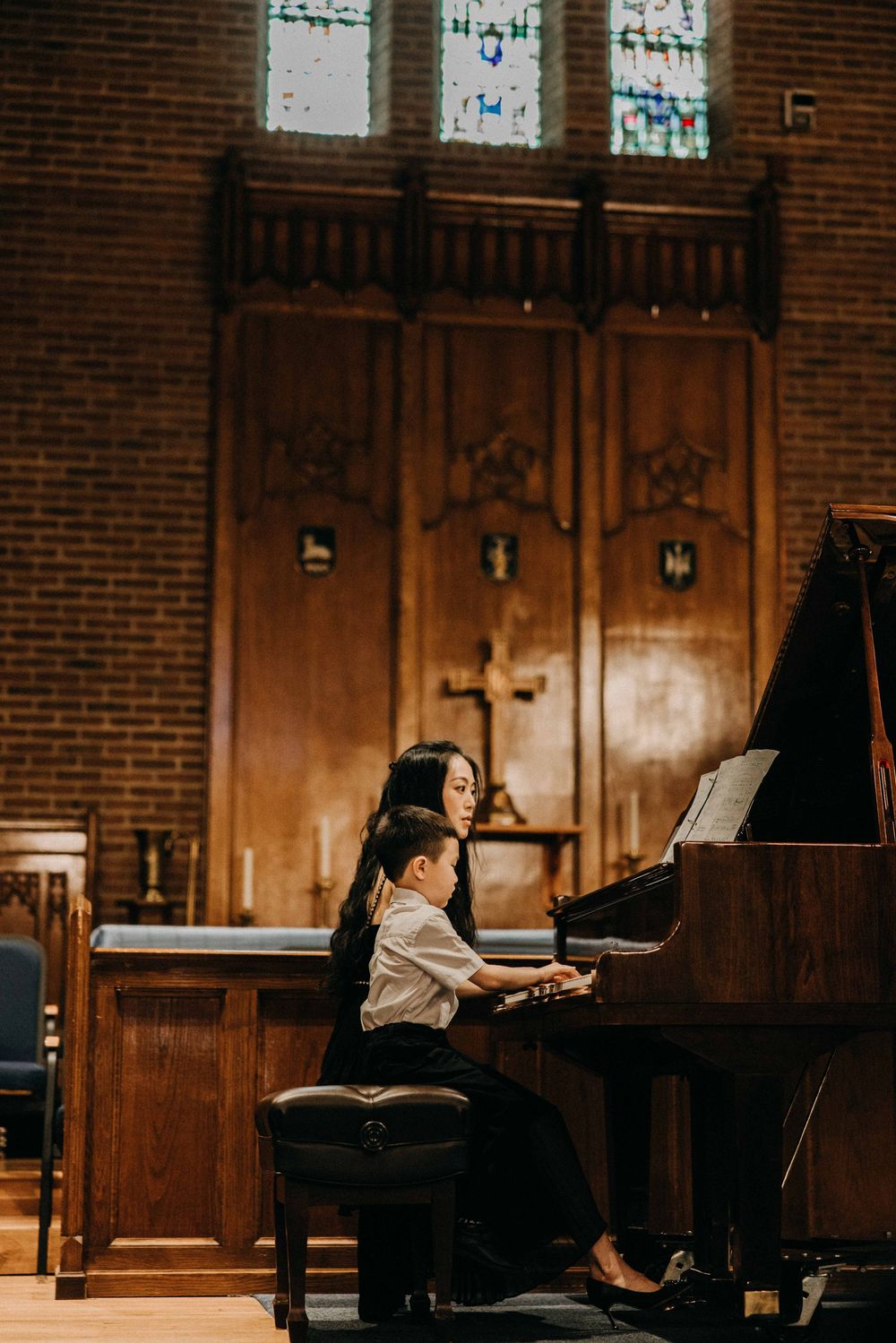 A woman and young boy play a grand piano together in an elegant church with stained glass windows.