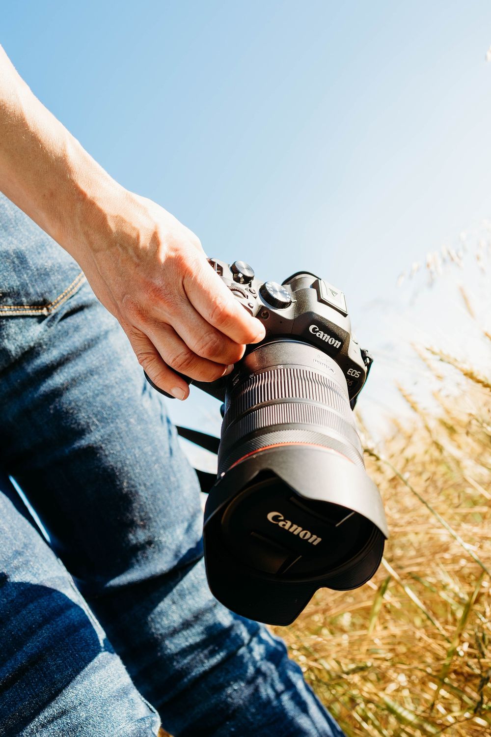 Person holding a Canon EOS DSLR camera with a large lens outdoors in a sunny field with blue sky.