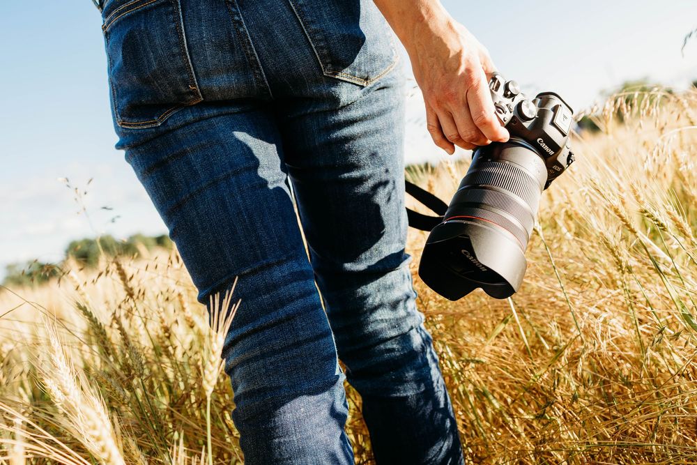 Photographer holding a DSLR camera walking through golden wheat field wearing ripped blue jeans on a sunny day.