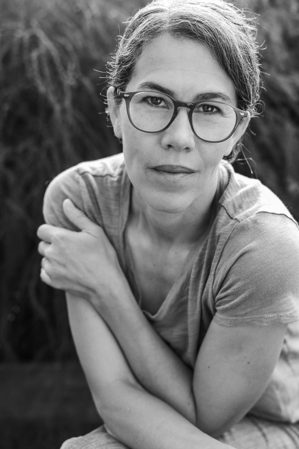 Black and white portrait of a woman with glasses and arms crossed, sitting outdoors with foliage background.