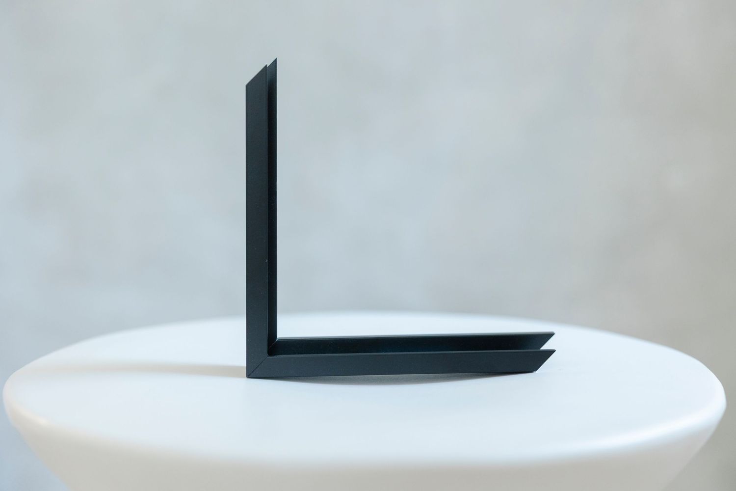 Black metal L-shaped bracket displayed upright on a white pedestal against a neutral gray background.