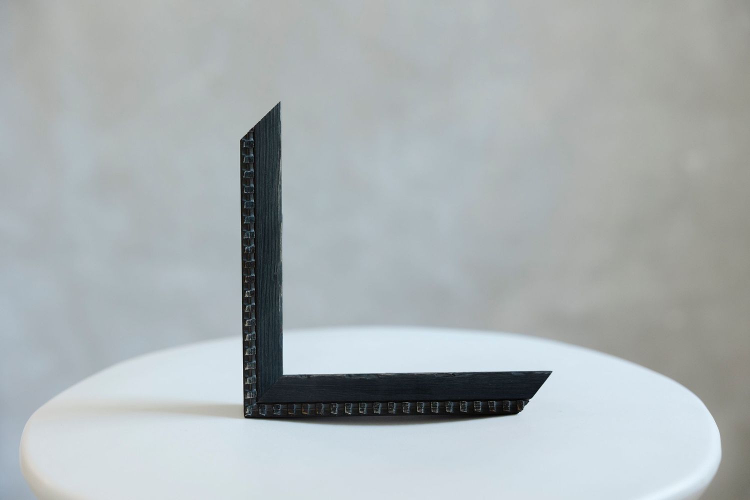 Black L-shaped metal angle bracket displayed upright on a white round table against a gray background.