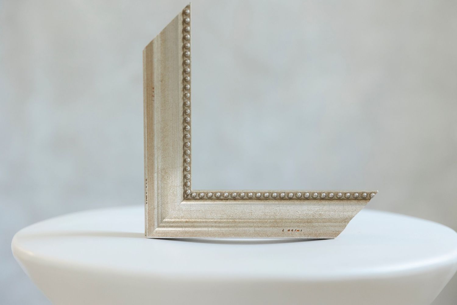 Silver ornate picture frame corner displayed on a white pedestal, featuring beaded detail trim and brushed metallic finish.