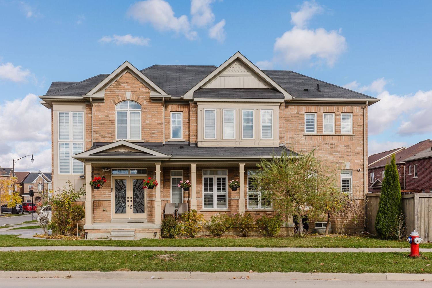 Two-story brick home with covered porch, manicured landscaping, and dark roof on a corner lot in a suburban neighborhood.