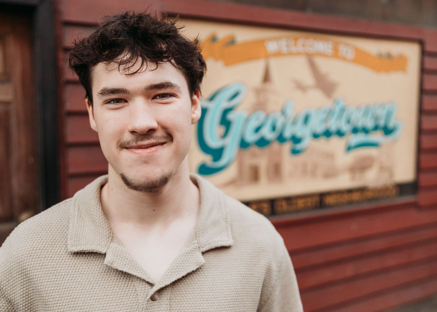 Young man smiling in front of a Welcome to Georgetown sign on a rustic wooden building.