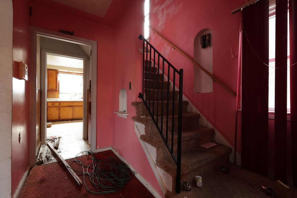 Dilapidated pink hallway with black iron staircase, peeling walls, and debris-covered floor in abandoned house.