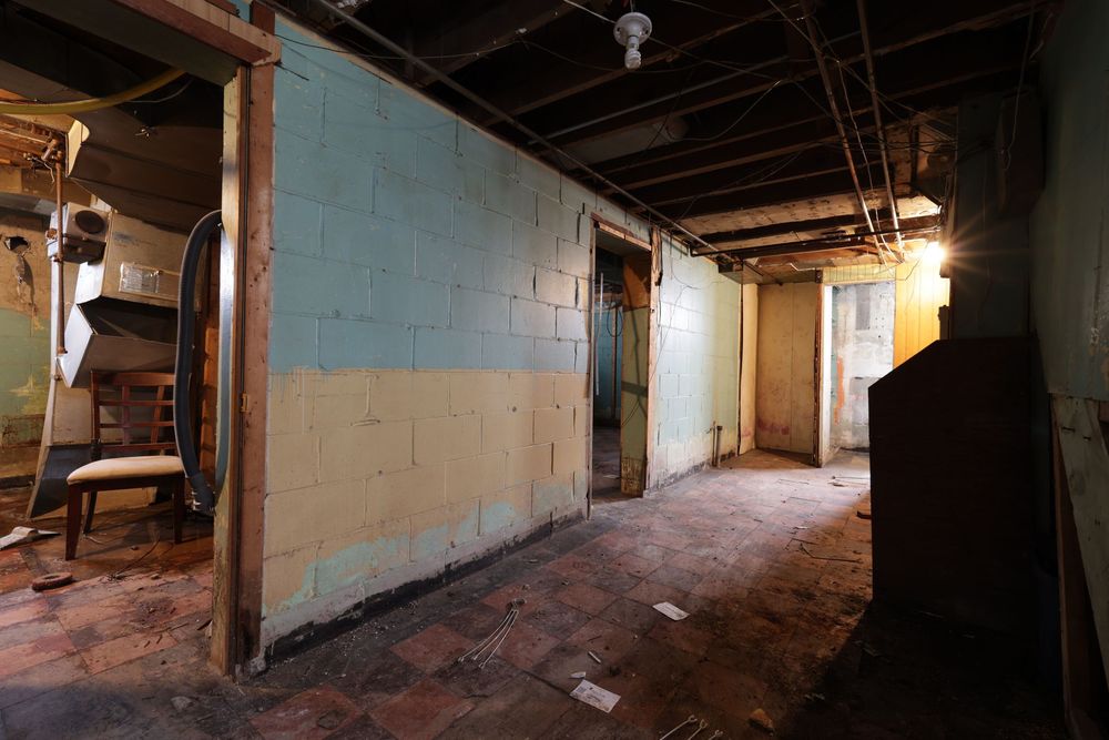Unfinished basement with concrete block walls, exposed ceiling, brick floor, and open doorways in a fixer-upper home.