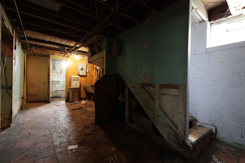 Dilapidated basement interior with peeling walls, exposed ceiling, debris on wet floor, and wooden staircase in poor condition.