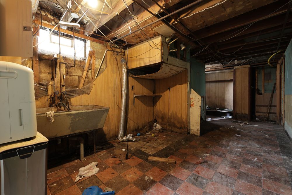 Abandoned basement with damaged walls, debris on tile floor, old sink, and exposed ceiling in a dilapidated building. Detroit Michigan