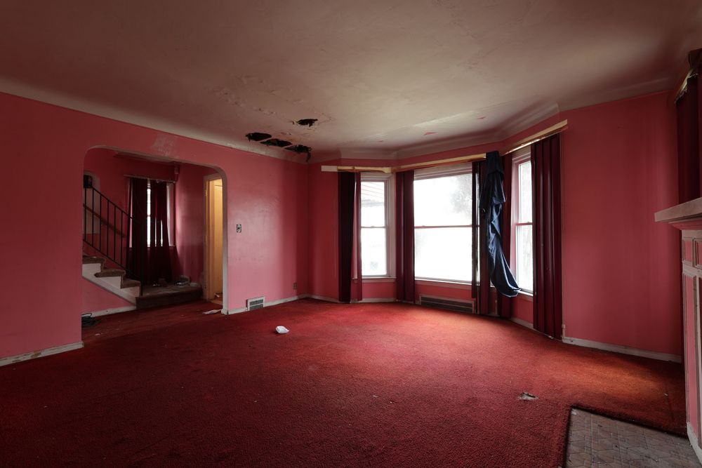 Empty living room with red carpet, pink walls, bay windows, and staircase visible in background, ceiling damage Detroit Michigan