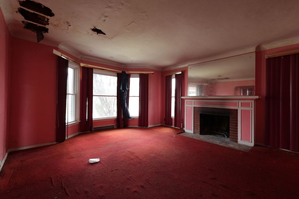 Abandoned room with red walls, red carpet, bay windows, and a white fireplace mantel in disrepair. ceiling damage Detroit Michigan