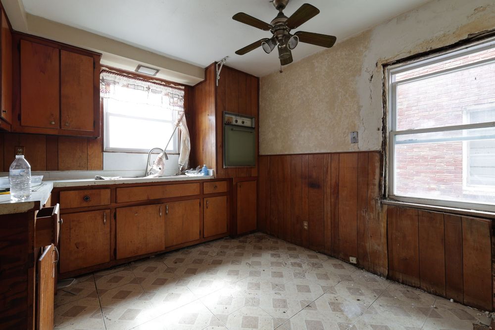 Outdated kitchen with wood cabinets, tile floor, ceiling fan, and natural light from two windows needing renovation.