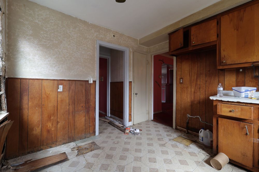 Neglected kitchen with wood-paneled walls, outdated cabinets, worn vinyl flooring, and debris in vacant home.