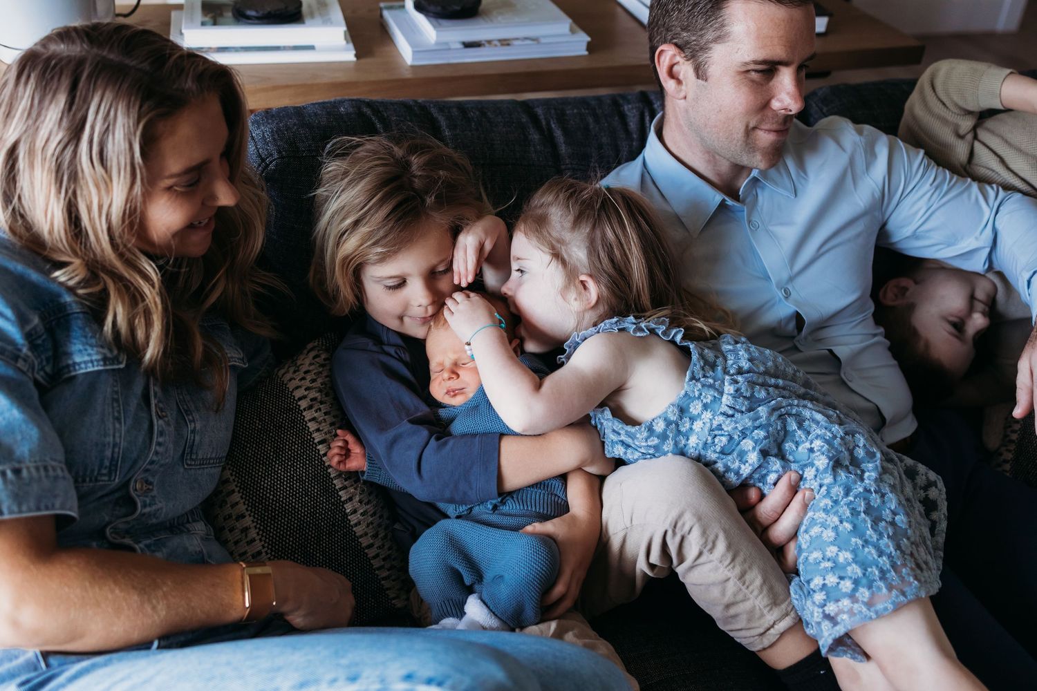 Family of four sitting on a couch, two young children sharing a tender moment while parents smile warmly nearby.
