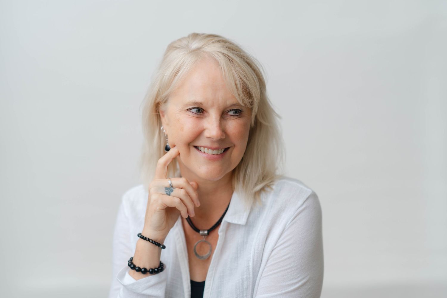 Smiling older woman with blonde hair wearing a white blouse and bracelet, posing confidently against a light background.