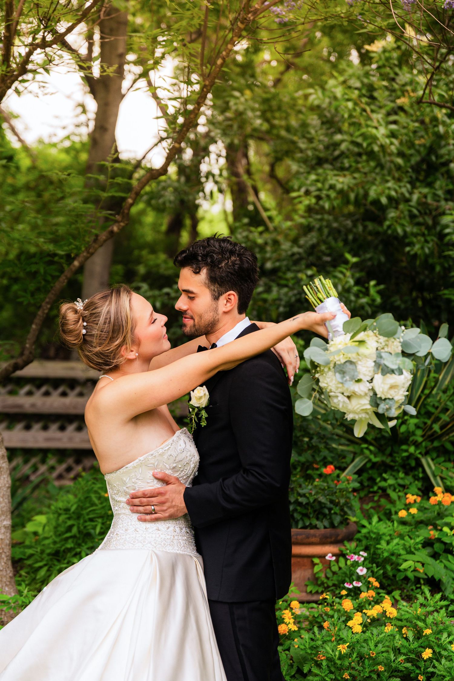 Bride and groom embrace in lush garden, she holds white floral bouquet, both smiling at each other in wedding attire.