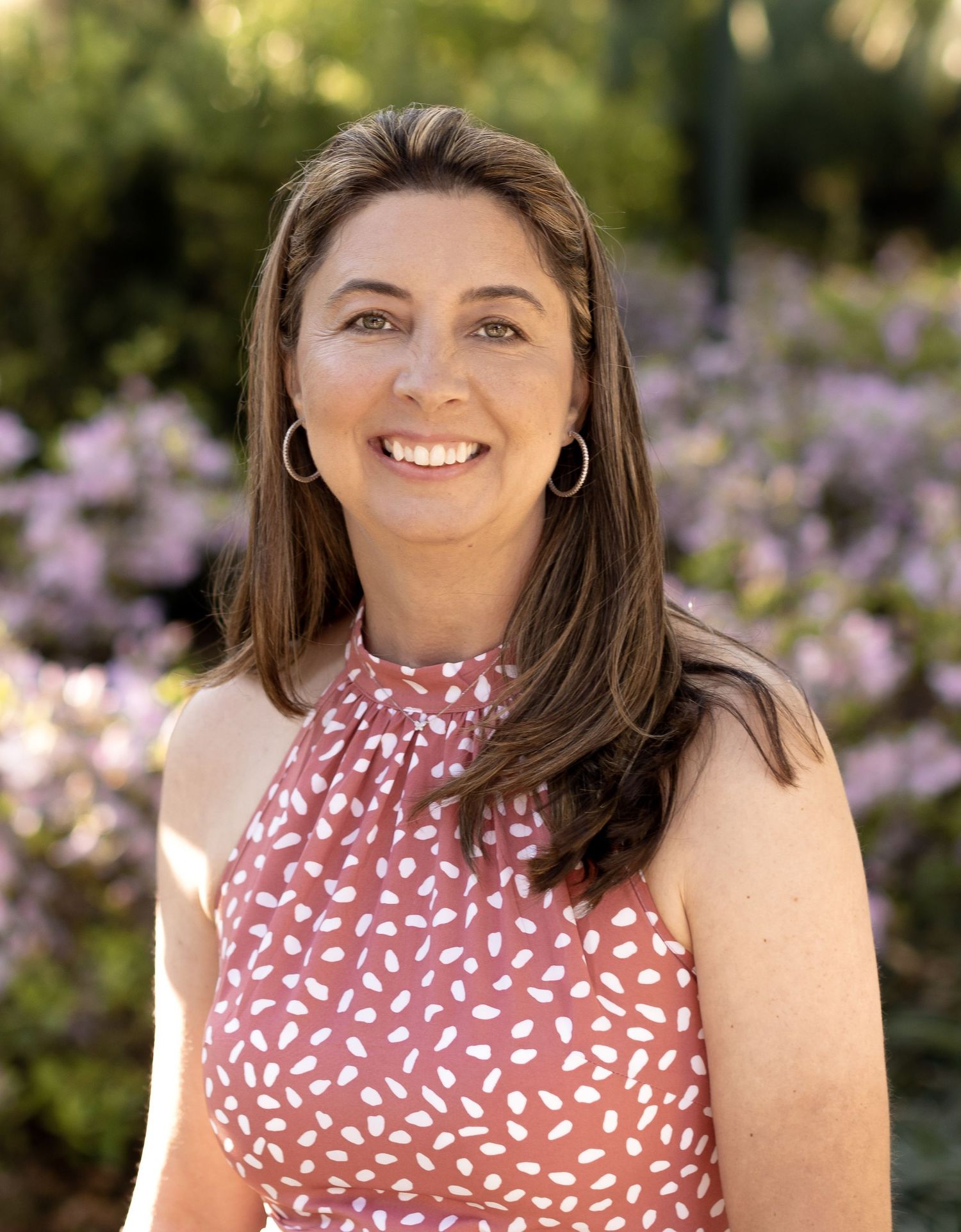 A person in a rust-colored polka dot halter top smiles warmly outdoors with purple flowers blooming in the background.