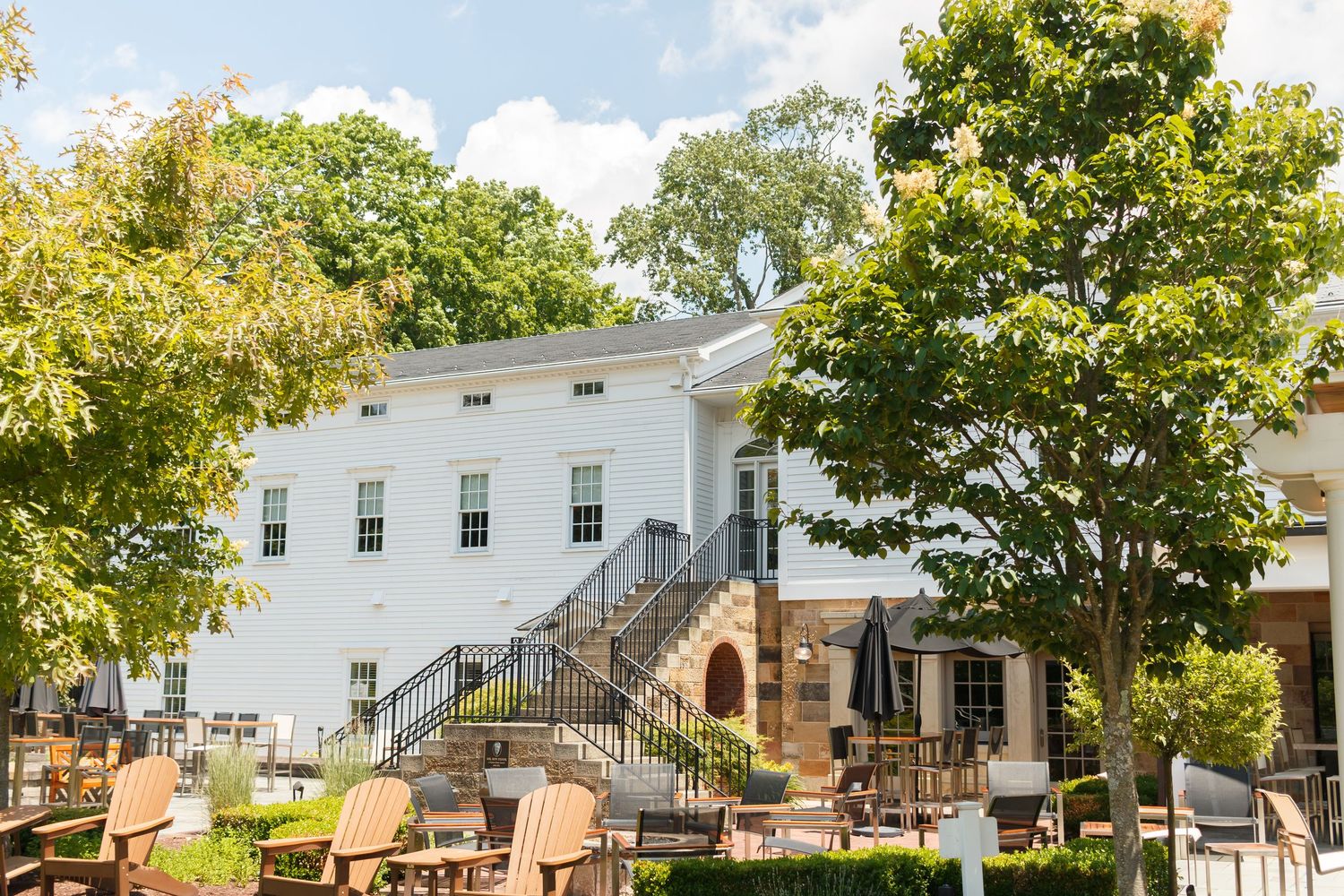 White colonial building with outdoor patio seating, Adirondack chairs, and lush green trees on a sunny day.
