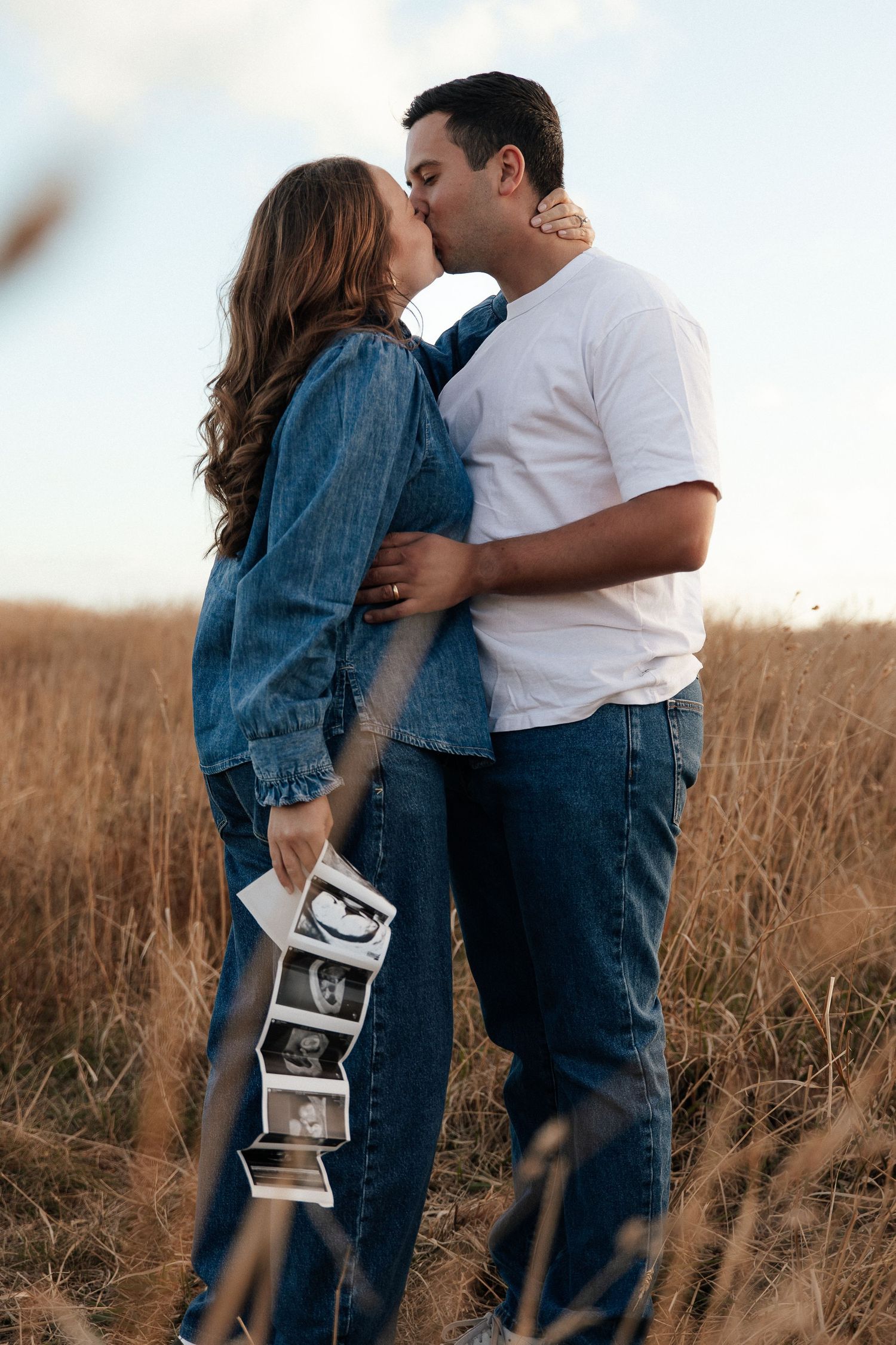 Couple kissing in golden field holding ultrasound photos, wearing denim, announcing pregnancy outdoors.