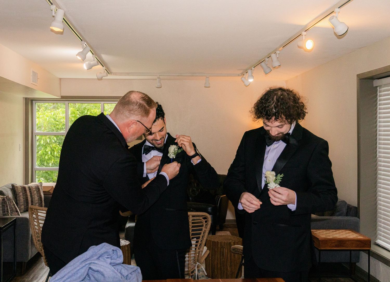 Three groomsmen in black suits pinning boutonnieres before a wedding ceremony indoors.