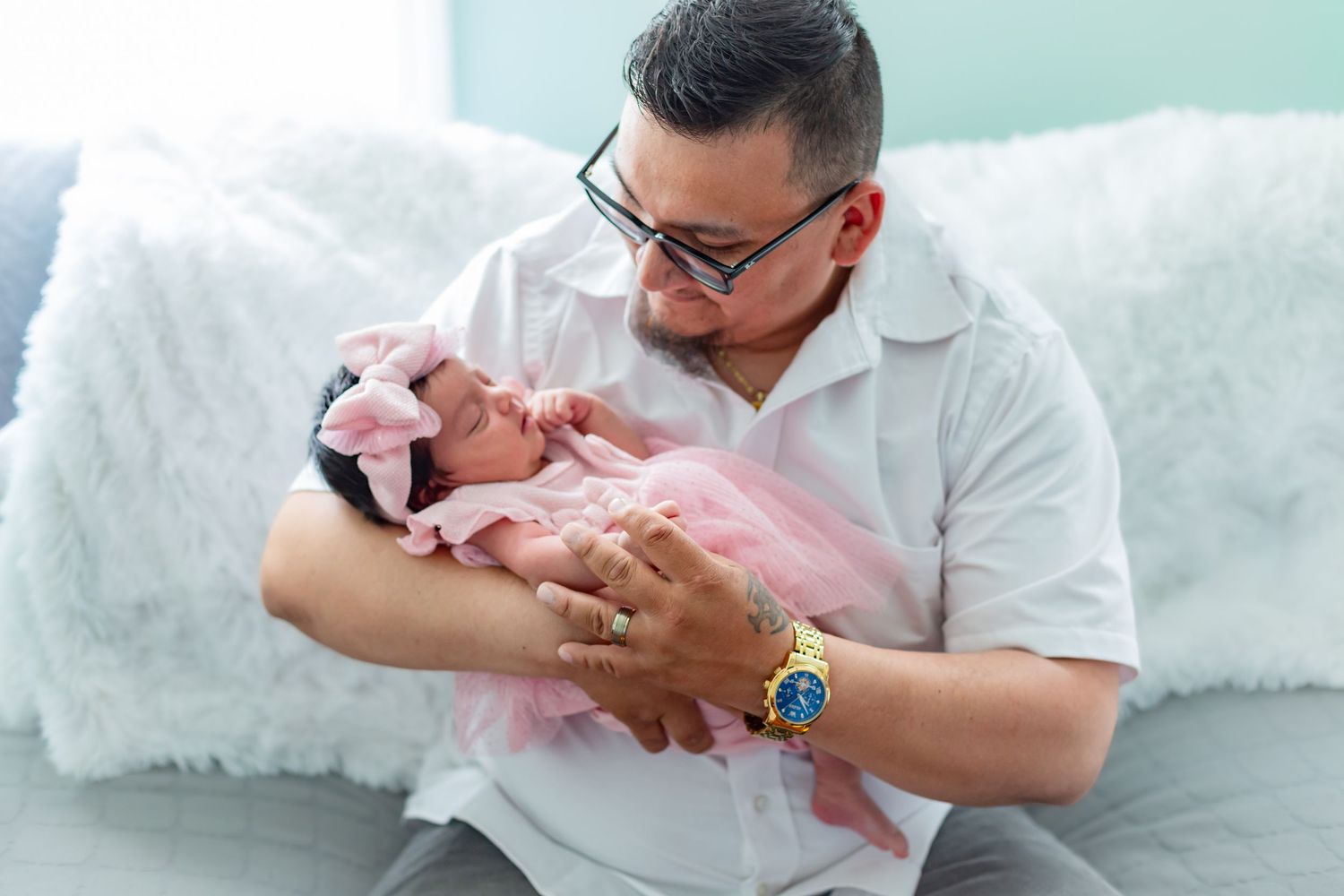Man in white shirt with gold watch tenderly holds sleeping newborn baby girl in pink tutu and bow headband.