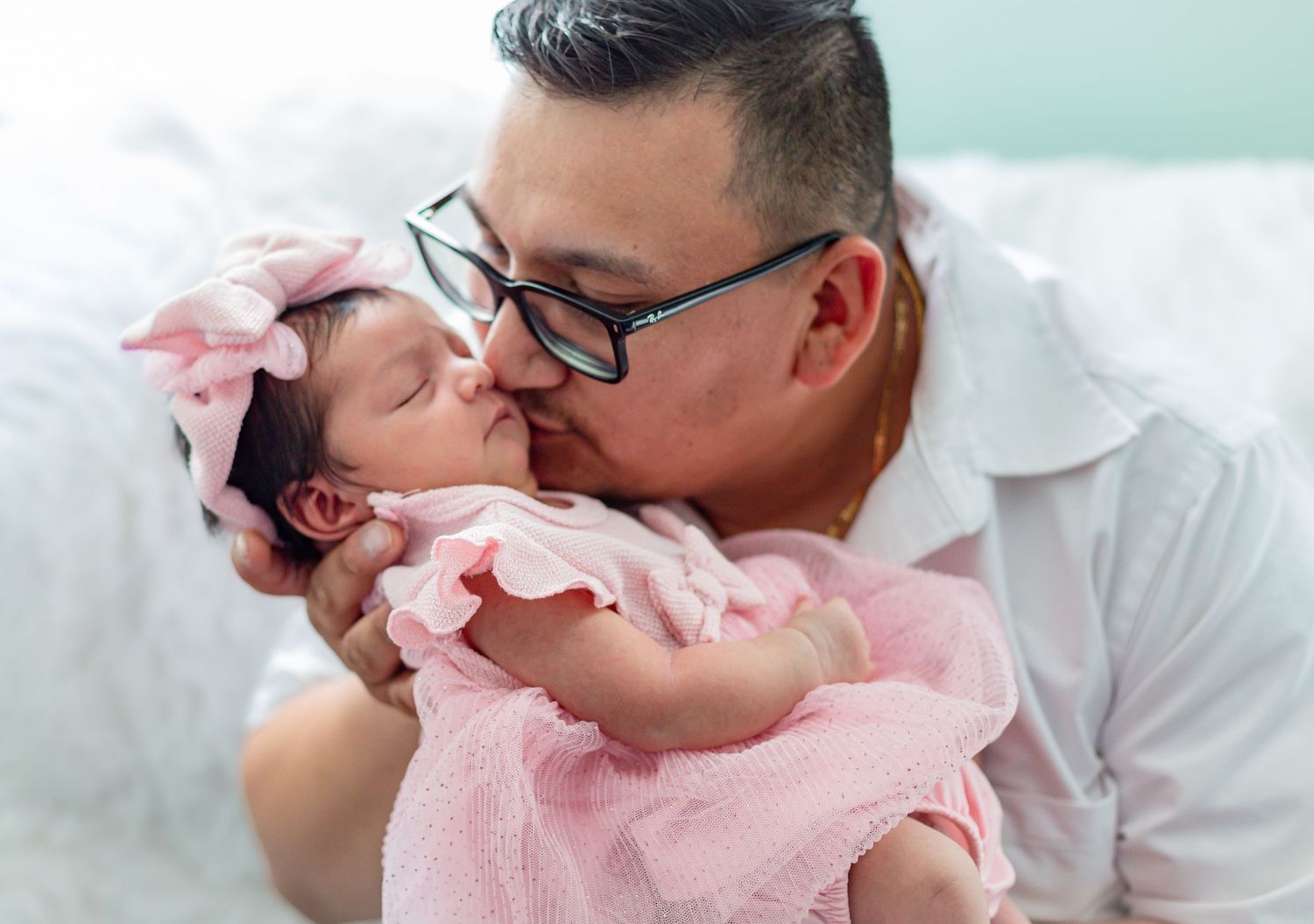 A father tenderly kisses his newborn baby girl dressed in pink with a bow headband.
