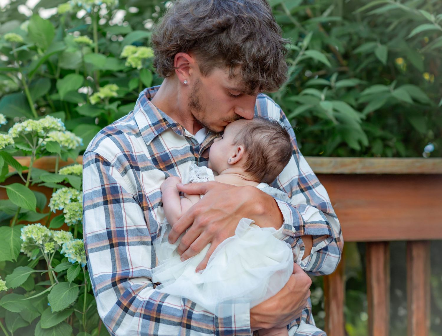 A young father in a plaid shirt tenderly kisses his newborn baby outdoors amid lush green foliage.