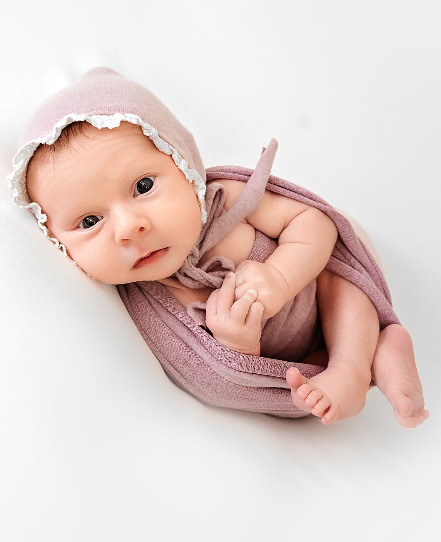 Adorable wide-eyed newborn baby peeking out from soft white blanket in black and white portrait.