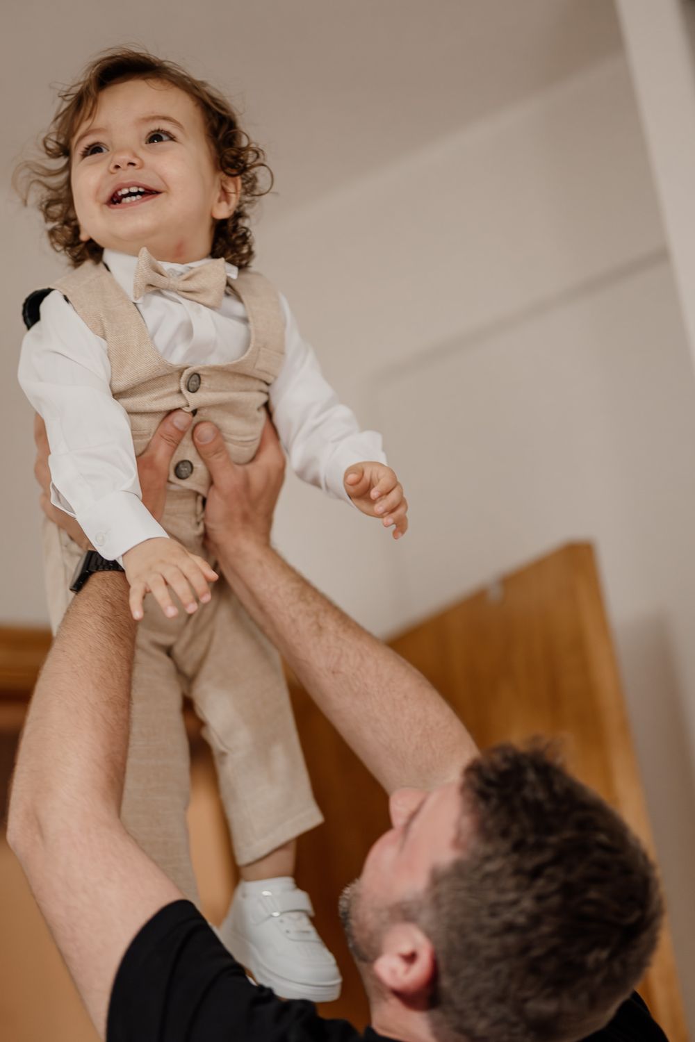 A joyful toddler in a beige suit lifted by his father, laughing with delight during a playful moment.