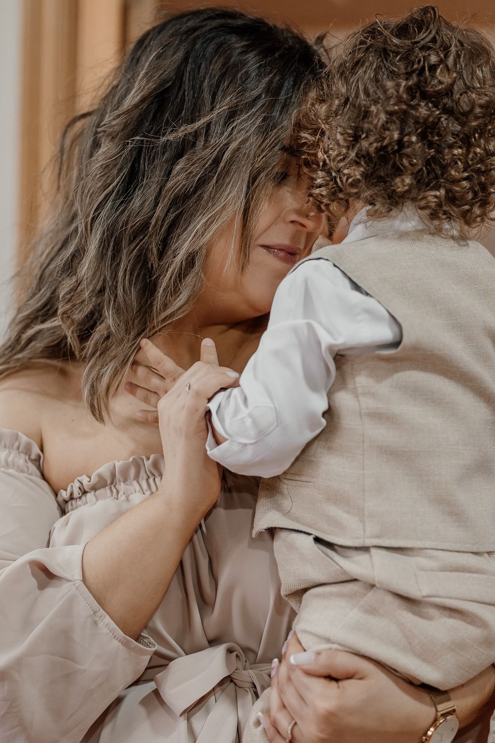 A woman in a beige off-shoulder dress tenderly embraces a curly-haired child in a linen suit.