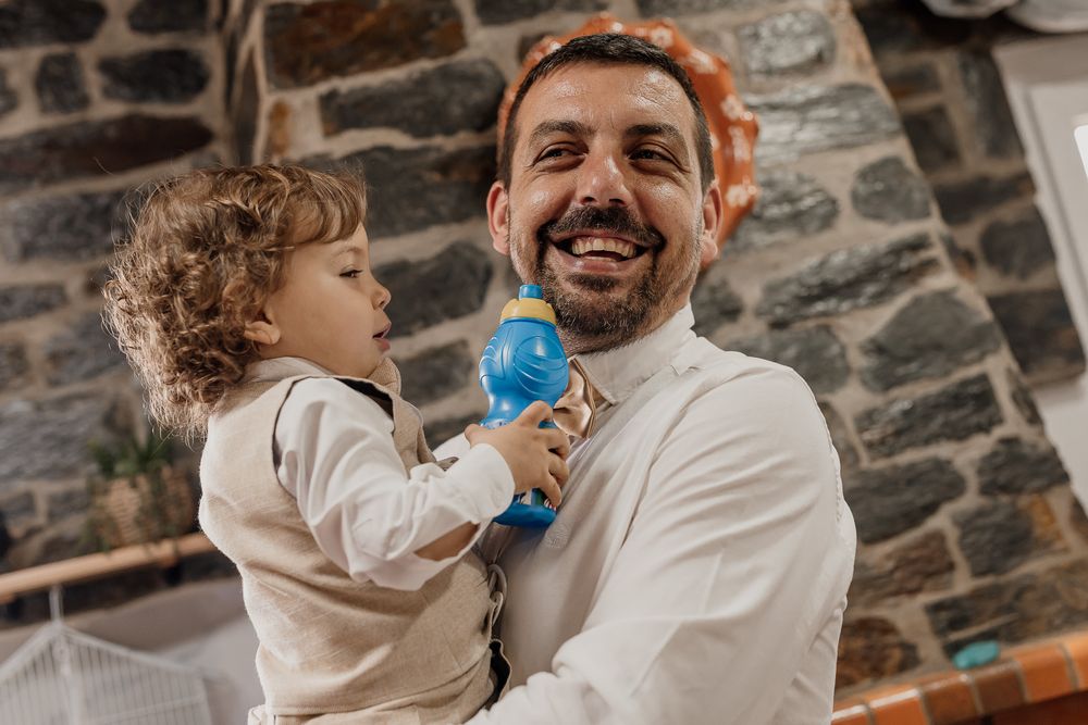 Smiling man holding a toddler with a blue toy at a rustic stone-walled venue.