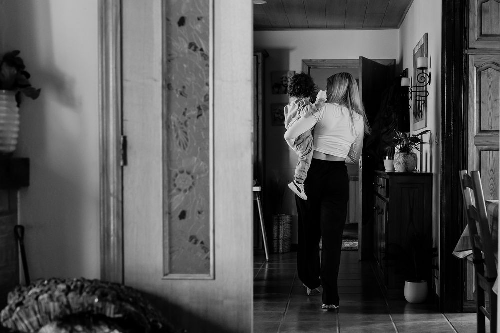 Black and white photo of a woman carrying a child through a hallway in a cozy home interior.