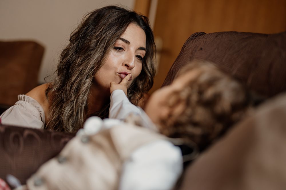 A woman with dark wavy hair gazes lovingly at a sleeping baby on a couch in a warm, cozy room.