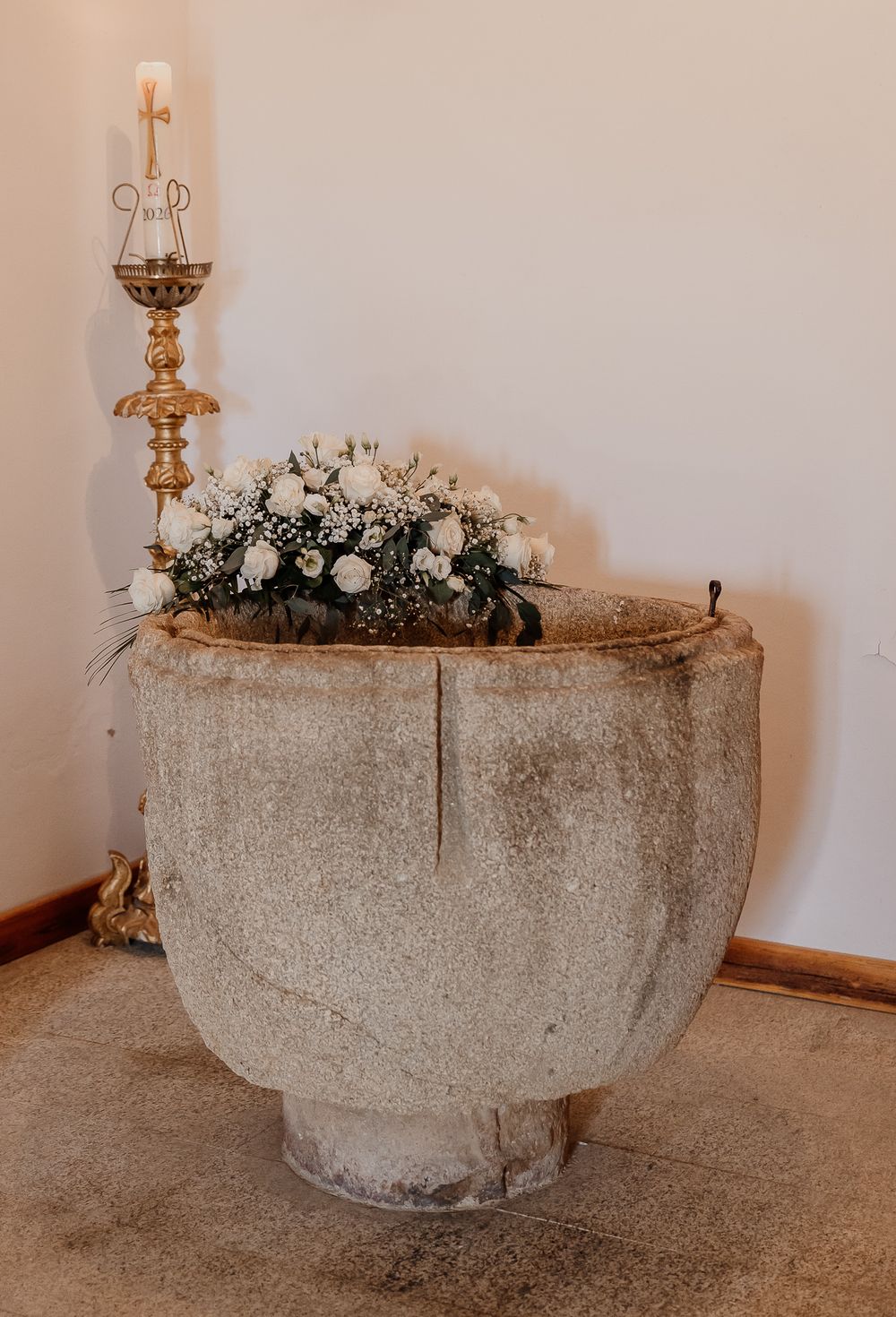 Ancient stone baptismal font decorated with white flowers and greenery beside a gold candlestick.