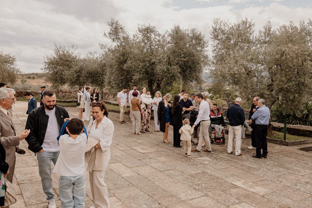 Wedding guests mingling outdoors near olive trees on stone paved courtyard on cloudy day.