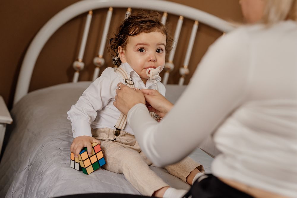 Toddler sitting on bed holding Rubik's cube while caregiver adjusts clothing in warm indoor setting.