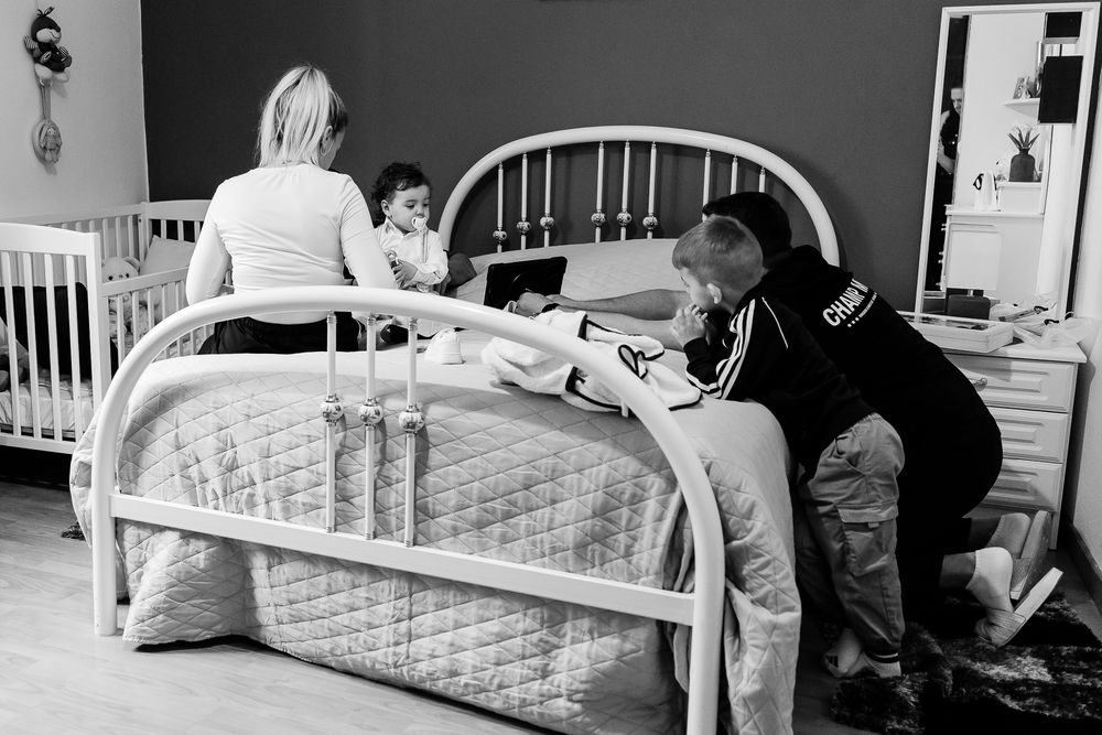 Black and white photo of a family with two children in a bedroom, parents tending to kids on a white bed near a crib.