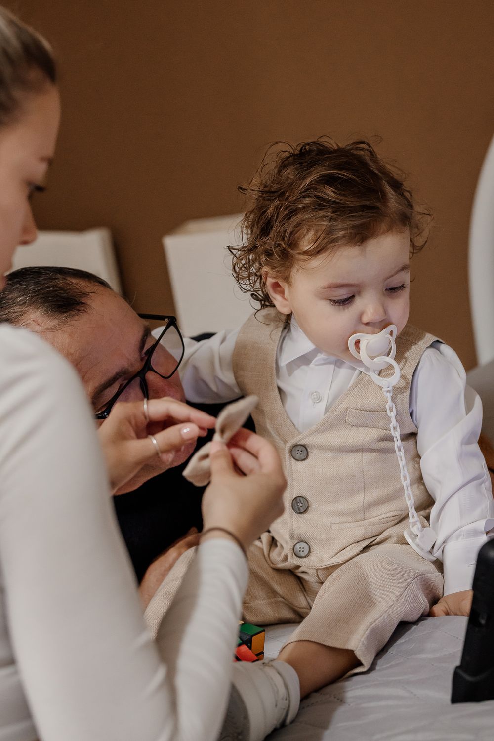 Toddler boy in beige vest and white shirt with pacifier being dressed by parents on bed.