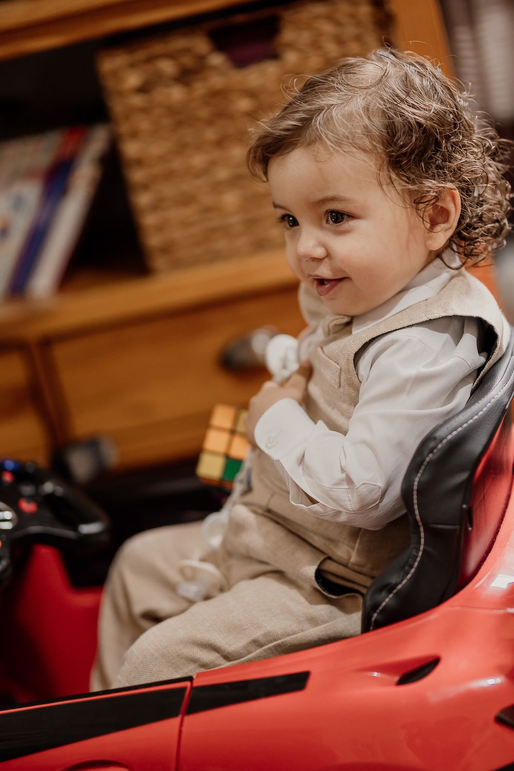 Smiling toddler with curly hair sitting in a red ride-on toy car, holding a small toy indoors.