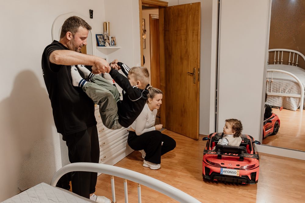 Family playing indoors, father lifting child while mother watches and toddler sits in red toy car.