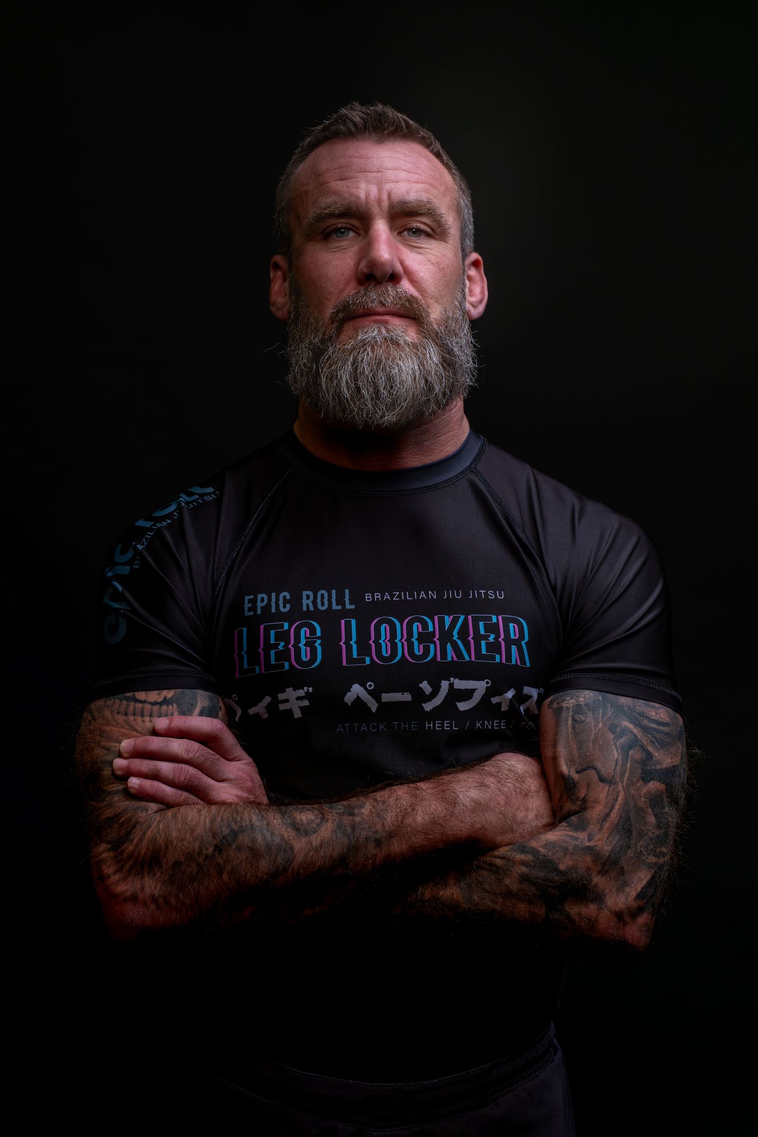Athletic man in black BJJ Locker t-shirt with gray beard and tattoos poses with crossed arms against dark background.
