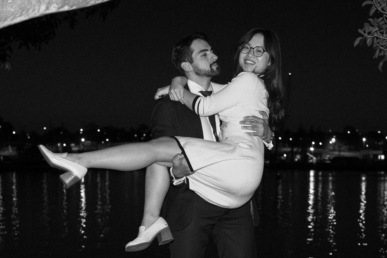 A couple in formal attire dances together at an evening waterfront venue with twinkling lights in the background.