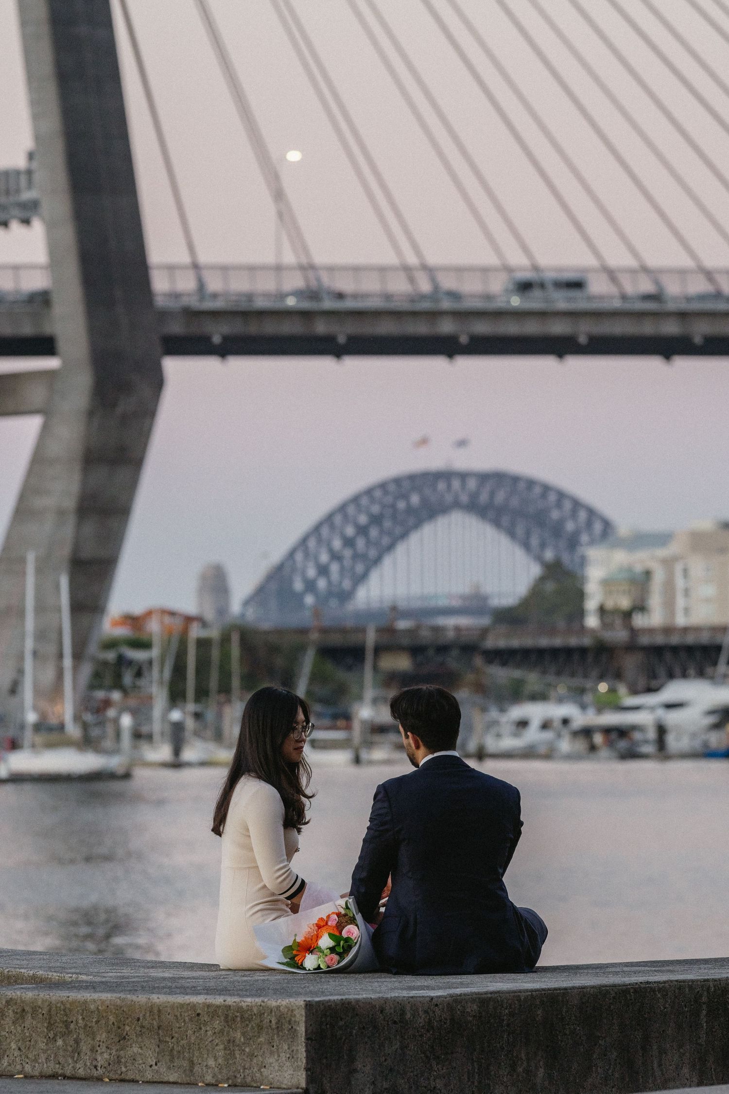 A couple sits by the waterfront beneath a large bridge on a cloudy day, creating a romantic urban scene.