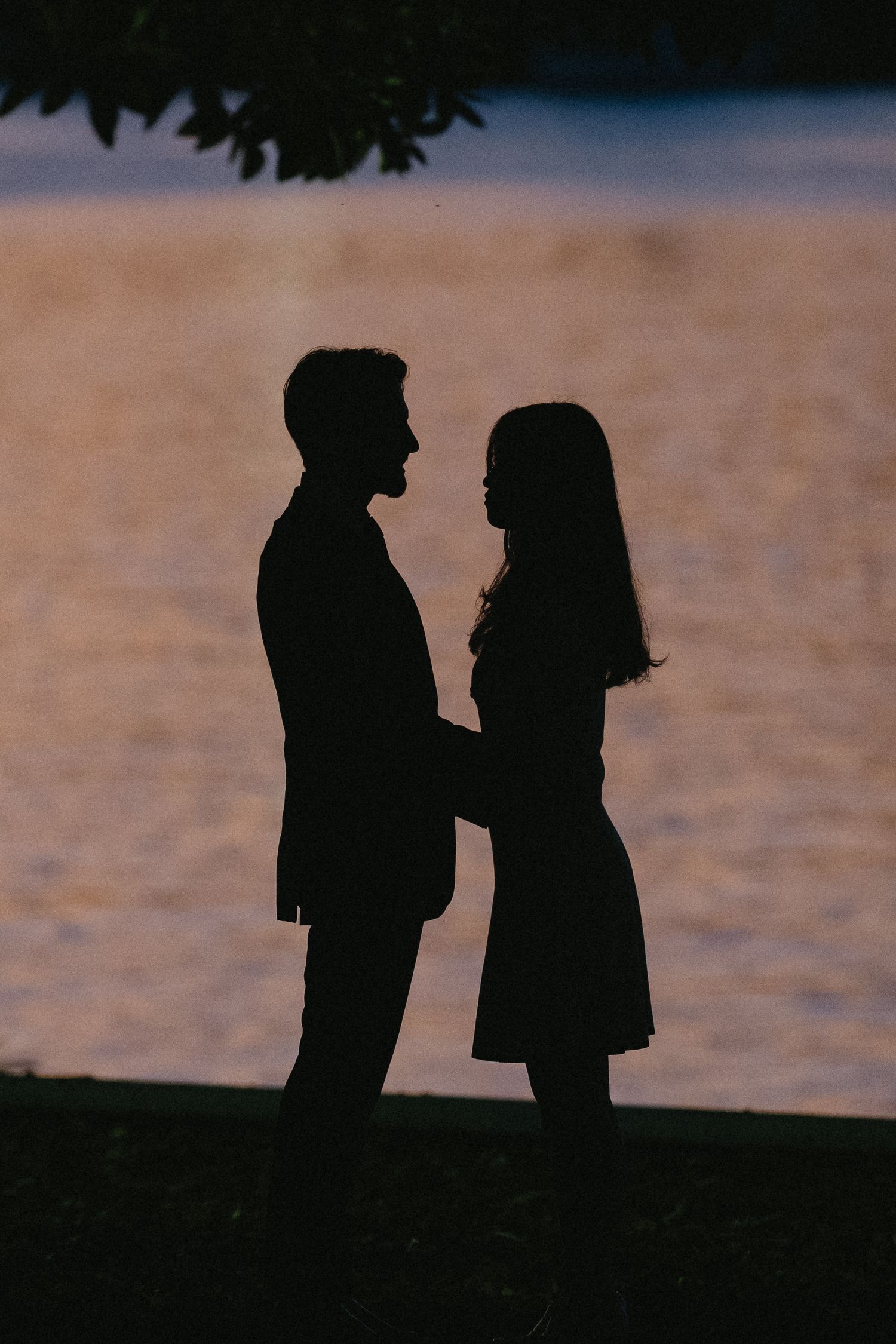 Silhouettes of two people standing together against a sunset sky reflected in calm water.